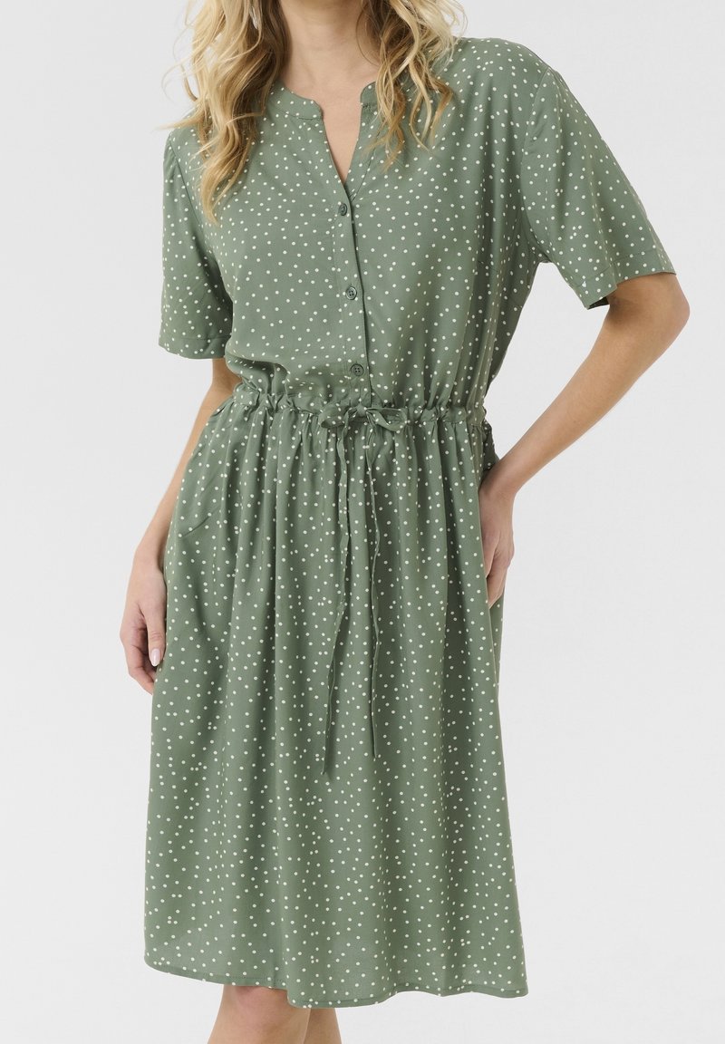Robe verte à manches courtes avec des pois blancs, boutons sur le devant et taille à cordon, portée par une personne avec la main sur la hanche.