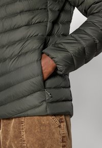 Osoba u zelenoj preklopnoj jakni s rukom u bočnoj džepu, uparena s smešnim kašmir hlačama protiv jednostavne pozadine.