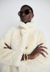 Kremfarvet faux fur frakke med høj krave, der har en knaplukning og struktureret stof. Sorte solbriller og metalliske armbånd fremhæver looket.