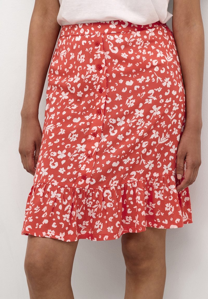Femme portant une jupe rouge à fleurs avec boutons sur le devant, un ourlet à volants et un haut blanc rentré, visible de la taille aux genoux.