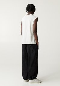 Camiseta blanca sin mangas con bordes sin rematar combinada con pantalones de mezclilla negros de corte amplio y zapatillas blancas, vista desde atrás.