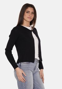 Cardigan nero realizzato in un tessuto testurizzato, caratterizzato da un colletto rotondo, maniche lunghe e un design cropped. Indossato sopra una camicia bianca.