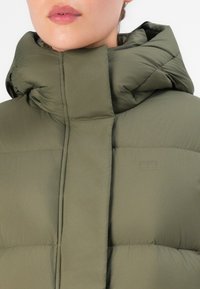Vrouw in een hoge, olijfgroene, gewatteerde winterjas met capuchon, gedeeltelijk dichtgeritst, waardoor alleen de onderkant van het gezicht en de nek zichtbaar zijn.