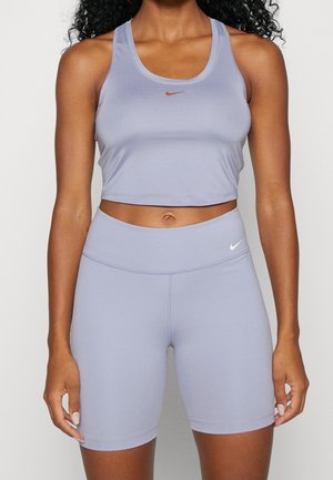 Femme portant un crop top Nike violet clair et un short de cycliste assorti, debout avec les bras relâchés le long du corps devant un fond neutre.