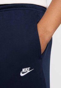 Navy Nike sporta bikses ar gludu audumu, sānu kabatām un baltu logotipa izšuvumu. Jostasvieta ir elastīga ērtam valkājumam.
