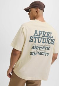 Tricou crem cu mânecă scurtă, având un graphic mare pe spate cu textul "APRIL STUDIOS" în albastru închis, însoțit de alte fraze.