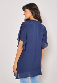 Marineblaue, locker sitzende Bluse mit fließenden Ärmeln, gerafftem Ausschnitt und transparenter Textur, die unterhalb der Hüften endet.