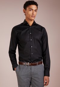 Chemise noire à manches longues avec une coupe slim, dotée d'un col classique, d'une fermeture à boutons et d'une texture lisse, associée à un pantalon à carreaux gris et une ceinture marron.