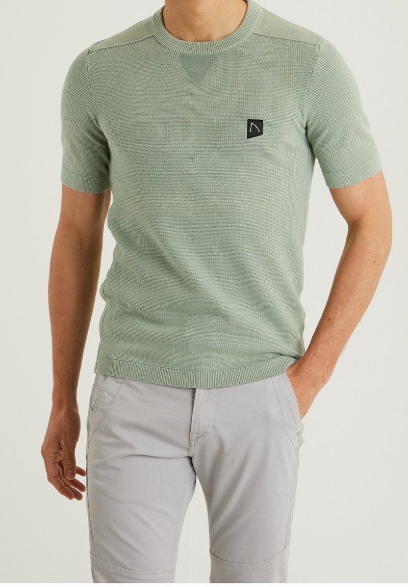 Mann trägt ein hellgrünes kurzärmeliges Strickshirt und hellgraue Hose, mit linker Hand in der Tasche und rechter Hand entspannt an der Seite.