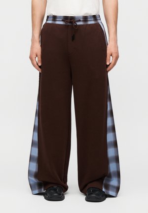 Άτομο που φορά καφέ παντελόνι με φαρδιά μπατζάκια, πλαϊνά πάνελ σε ανοιχτό μπλε και μαύρο καρό και μαύρα παπούτσια, στέκεται σε λευκό φόντο.