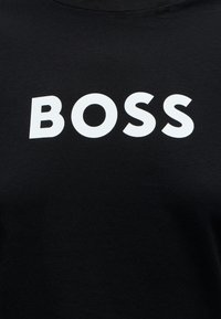 Sort stof med fede hvide store bogstaver, der danner ordet "BOSS", centreret på brystområdet af et beklædningsgenstand.