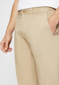 Pantalons beiges en tissu lisse, dotés d'une fermeture à bouton et de poches latérales, avec une coupe droite et un design minimaliste.