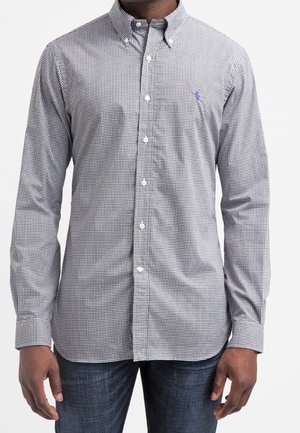 Homme portant une chemise à manches longues à carreaux noirs et blancs boutonnée avec un petit logo sur la poitrine, associée à un jean bleu foncé.