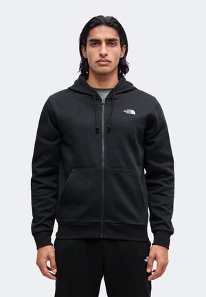 Homme aux cheveux bruns mi-longs portant un sweat à capuche noir zippé et un pantalon assorti avec un logo blanc sur la poitrine et la cuisse, debout face à l’avant.