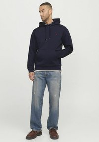 Sweatshirt à capuche bleu marine en matière douce, avec une poche avant et un petit logo rouge. Porté avec un jean délavé et des chaussures brunes.