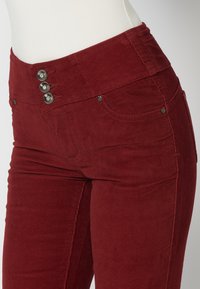 Pantalon en velours côtelé rouge taille haute avec trois boutons métalliques décoratifs et des poches avant sur un mannequin blanc.