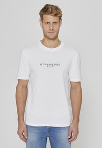 T-shirt en coton blanc à manches courtes et col rond, avec l'inscription "STRENESSE BLUE" en noir sur la poitrine. Porté avec un jean bleu.