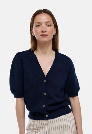 Femme aux cheveux bruns lisses porte un cardigan bleu marine texturé à manches courtes avec des boutons et un pantalon clair à rayures verticales.