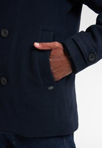 Cappotto in misto lana blu navy con tasca laterale, chiusure a bottoni e polsini testurizzati, con un dettaglio logo metallico sulla cucitura della tasca inferiore.