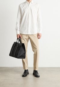Borsa tote in pelle nera testurizzata con doppi manici, indossata con una camicia bianca abbottonata e pantaloni beige, completata da scarpe nere.
