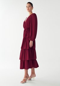 Burgundowa, warstwowa maxi sukienka z długimi rękawami, ściśniętą talią i głębokim dekoltem w kształcie litery V. Modelka zestawiona z białymi sandałami na obcasie. Gładka tekstura materiału.