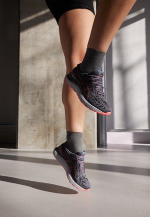 Piernas con calcetines grises y zapatillas de correr negro-rosas saltando en interiores con la luz del sol proyectando sombras en el suelo y la pared.