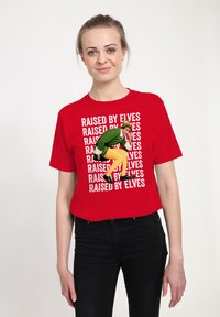 Červené tričko s krátkým rukávem s opakujícím se bílým textem "RAISED BY ELVES" a ilustrací zeleného skřítka v hravé pozici na přední straně.