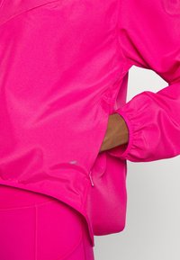 Jasnoróżowa, lekka kurtka o luźnym kroju, z marszczonymi mankietami i boczną kieszenią. Gładka tekstura z delikatnym połyskiem.