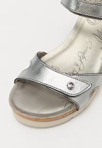 Silberne metallische Sandale mit zwei verstellbaren Riemen, weichem Fußbett und flacher Sohle. Verfügt über einen kleinen dekorativen Knopf am Riemen.