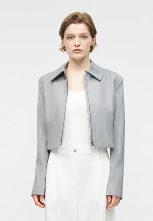 Γυναίκα με κοντά μαλλιά που φοράει ανοιχτό γκρι κοντό μπουφάν πάνω από λευκή μπλούζα και ψηλόμεσα λευκά παντελόνια, στραμμένη προς τα εμπρός με φόντο απλό.