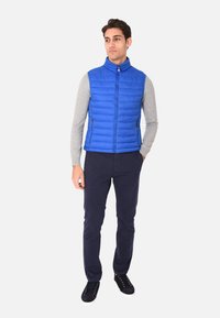 Gilet blu gonfiato con cuciture verticali, colletto alto e due tasche laterali con zip, indossato sopra una maglietta grigia a maniche lunghe e pantaloni blu navy.