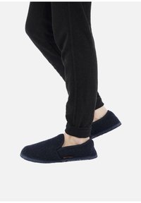Persona che indossa pantaloni neri e pantofole slip-on di un blu scuro, con un piede leggermente sollevato, su uno sfondo bianco uniforme.