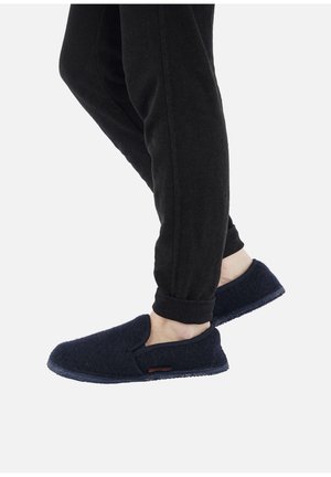Person wearing black pants and dark navy slip-on slippers, one foot slightly raised, against a plain white background.