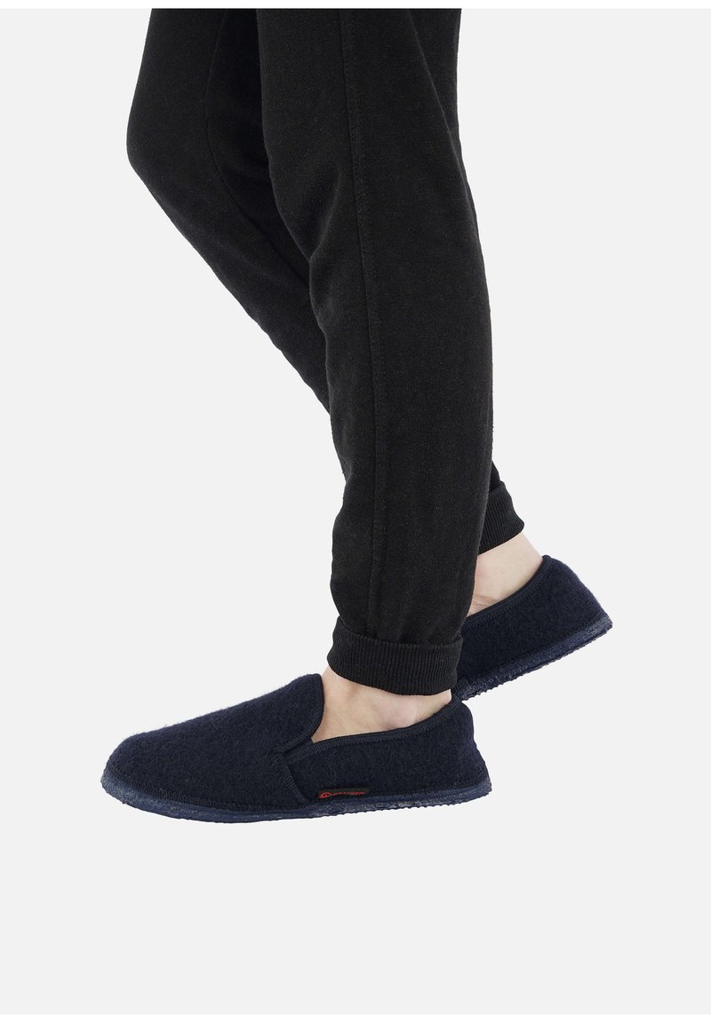 Persona che indossa pantaloni neri e pantofole slip-on di un blu scuro, con un piede leggermente sollevato, su uno sfondo bianco uniforme.