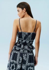 Sort peplum kjole med hvide blomsterdesign, tynde justerbare stropper og flæser, vist fra ryggen mod en lyseblå baggrund.