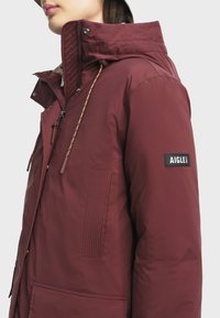 Bordeauxkleurige geïsoleerde jas met hoge kraag, ritsluiting en een gebreide textuuraccent. Bevat een logo patch op de mouw.