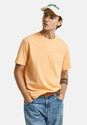 Oranges Baumwoll-T-Shirt mit kurzen Ärmeln und Rundhalsausschnitt; verfügt über ein kleines weißes Logo auf der Brust. Getragen mit blauen Jeans und einem braunen Gürtel.