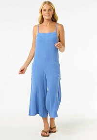 Mono azul sin mangas con tirantes ajustables, tejido texturizado, piernas amplias y dos bolsillos frontales. Llevado con sandalias marrones.