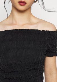 Svart off-shoulder topp gjord av texturerad tyg med rynkade detaljer och subtil lyster, med korta, volangärmar och en tajt passform.
