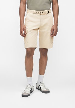 Άνδρας που φοράει μπεζ cargo σορτς με ζώνη, λευκές κάλτσες και λευκά αθλητικά παπούτσια με μαύρες ρίγες, στέκεται μπροστά σε απλό φόντο.