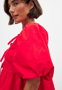 Robe en coton rouge avec des manches bouffantes volumineuses, des accents de nouage au niveau du décolleté et une jupe froncée. Texture lisse et couleur vive.