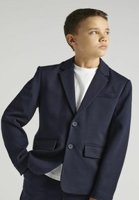 Blazer navy avec un col à revers, deux poches avant et deux boutons, fabriqué en tissu lisse, porté sur un T-shirt blanc.