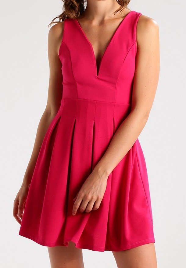 Femme portant une robe rose vif sans manches avec un profond décolleté en V et une jupe plissée sur un fond gris uni.