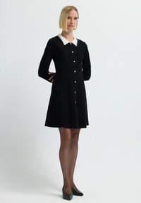 Vestito nero con maniche lunghe, colletto bianco, chiusura a bottoni e gonna plissettata. Il tessuto ha una superficie liscia, con sottili righe verticali.