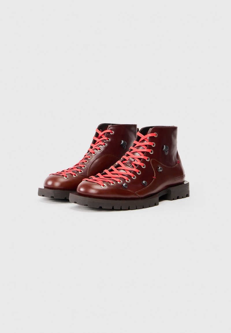 Ein Paar glänzende, dunkelbraune Lederstiefel mit leuchtend roten Schnürsenkeln und dicken schwarzen Sohlen auf einem schlichten hellgrauen Hintergrund.