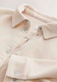 Maglia in pile beige con superficie testurizzata, bottoni a pressione in metallo e colletto. Lo strato interno è un tessuto beige liscio.