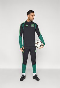 Fekete sportos melegítő zöld díszítéssel, cipzáras felsővel, hálós ujjakkal és hozzá illő nadrággal. A modell futballlabdát tart.