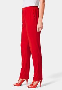 Pantalons rouges à taille élastique avec plis à l'avant, jambes droites et texture lisse. Associés à des chaussures à talons ouverts rouges.