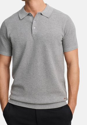 Homme portant un polo en maille à manches courtes gris clair avec une patte à deux boutons et un pantalon noir, les mains dans les poches.