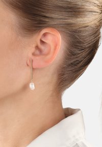 Orecchini in oro con un design a barra lunga e sottile, caratterizzati da un'unica goccia di perla bianca irregolare che pende elegantemente vicino al lobo dell'orecchio.